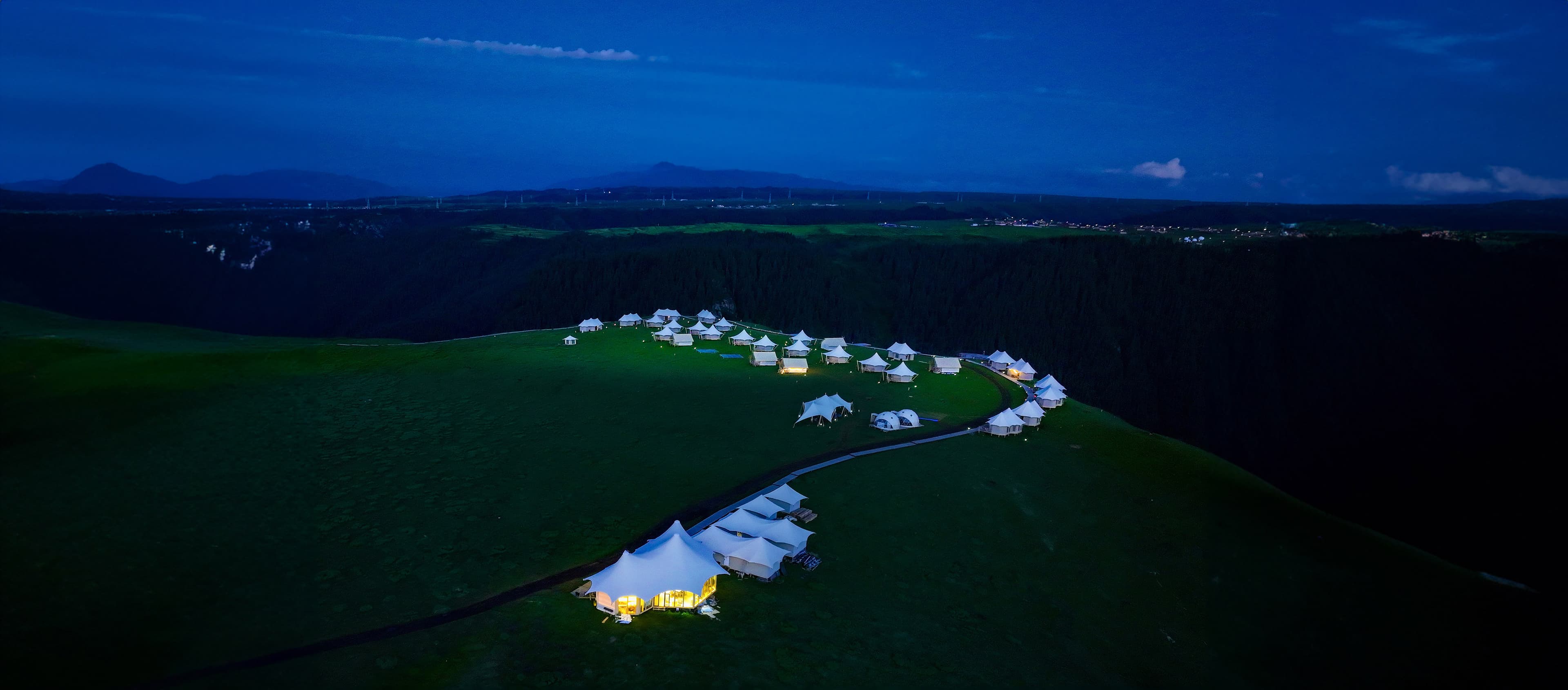 Nighttime aerial view of glowing white tents scattered across a lush green mountain plateau.