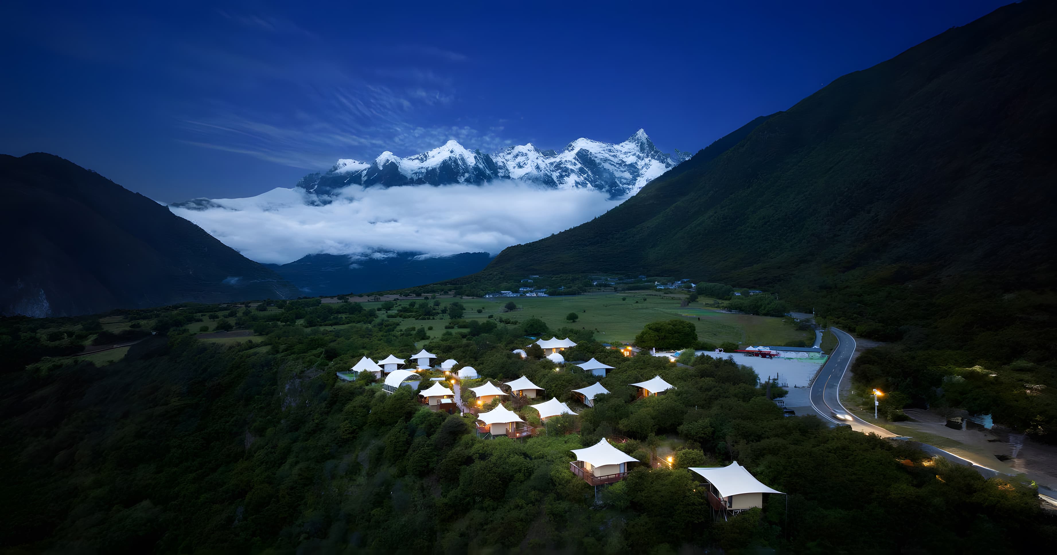 Glowing white tents on a lush hillside beneath majestic snow-capped mountains at dusk.