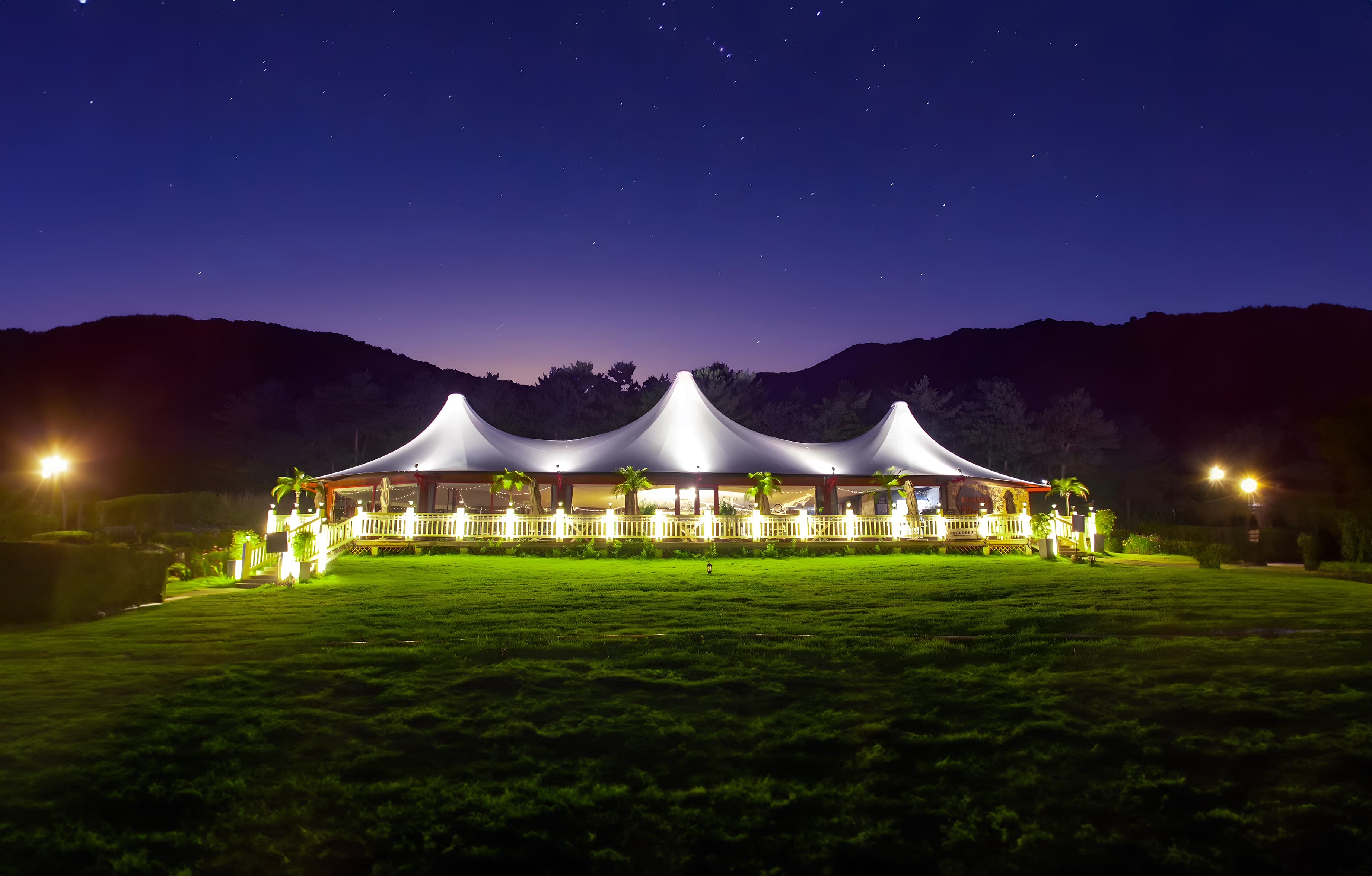Illuminated white marquee on a green lawn under a starry sky with dark mountains.