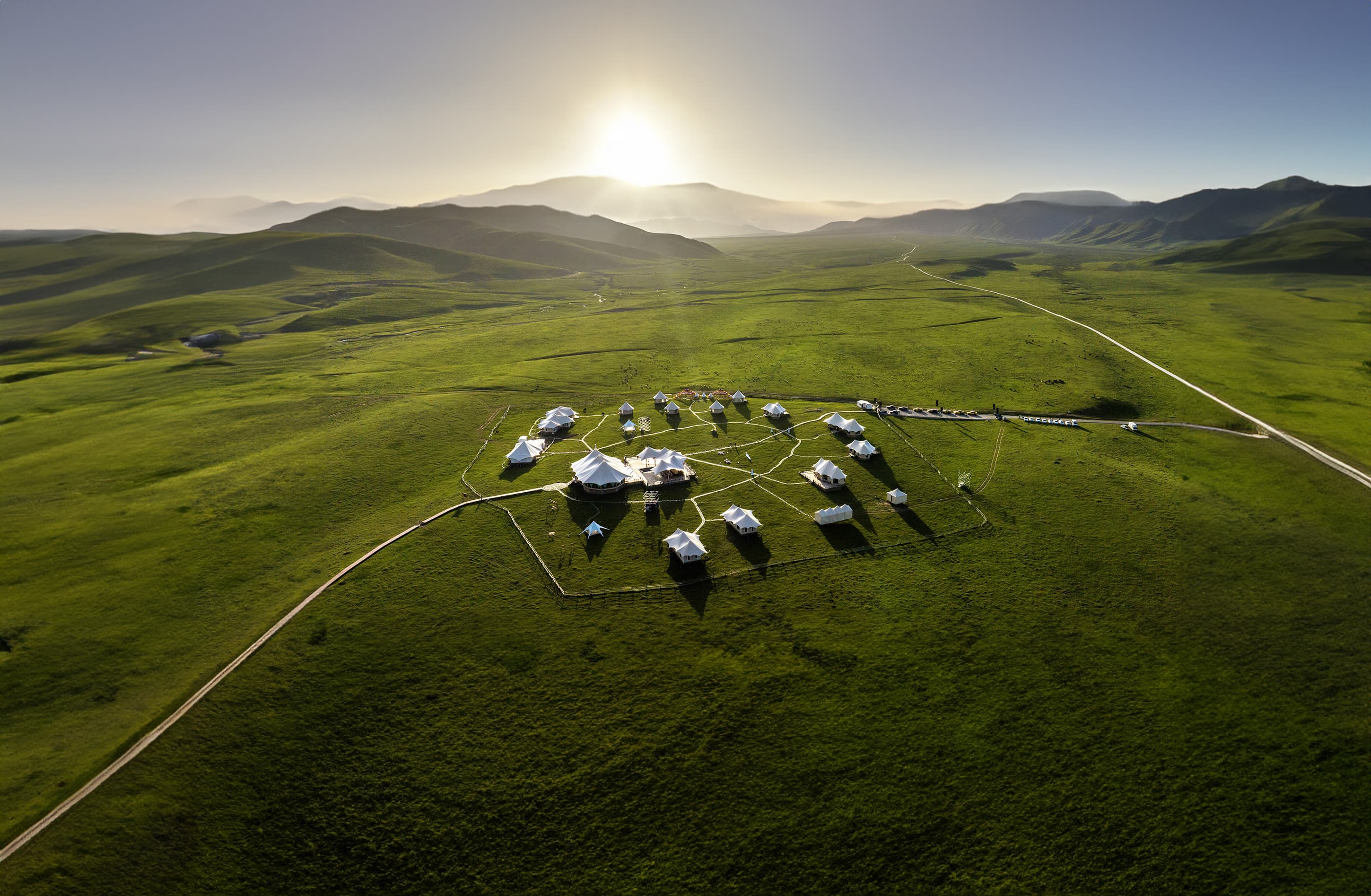 Aerial view of white glamping tents in a vast green valley during a bright sunrise.