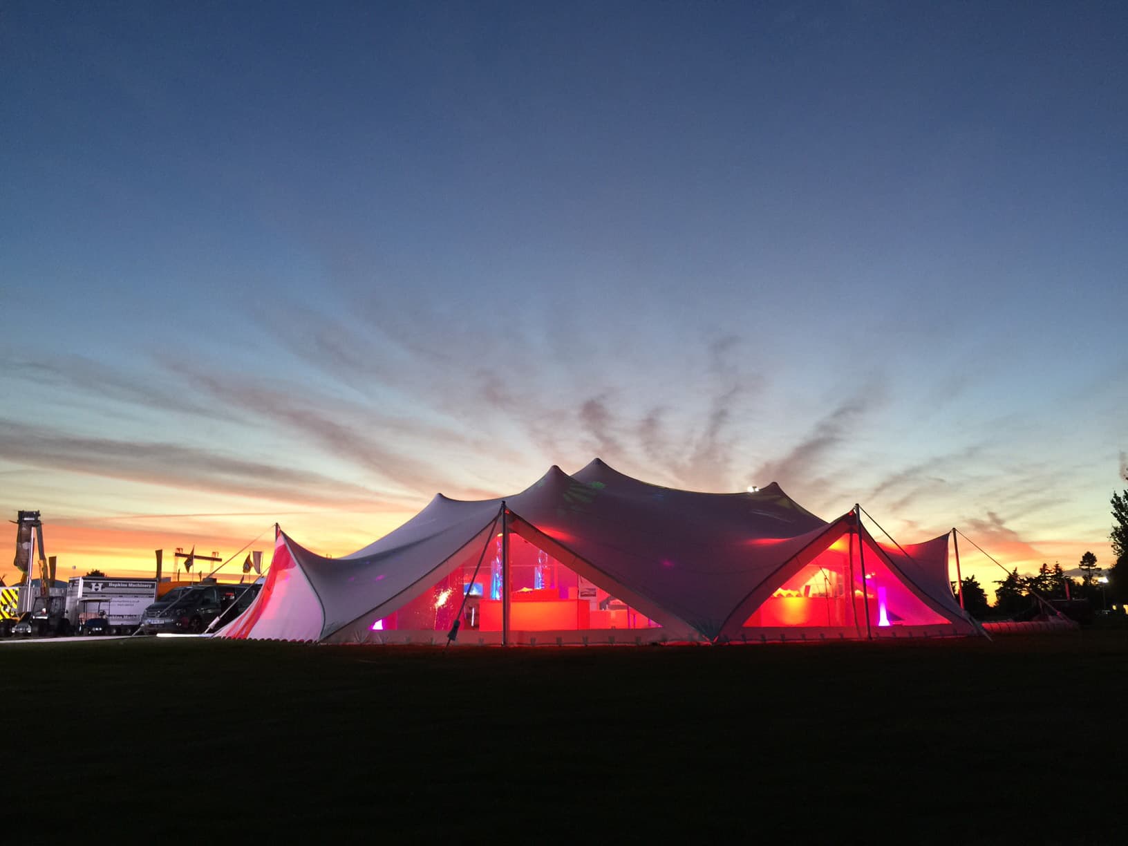 Large white event tent glowing with red light under a dramatic sunset sky at dusk.