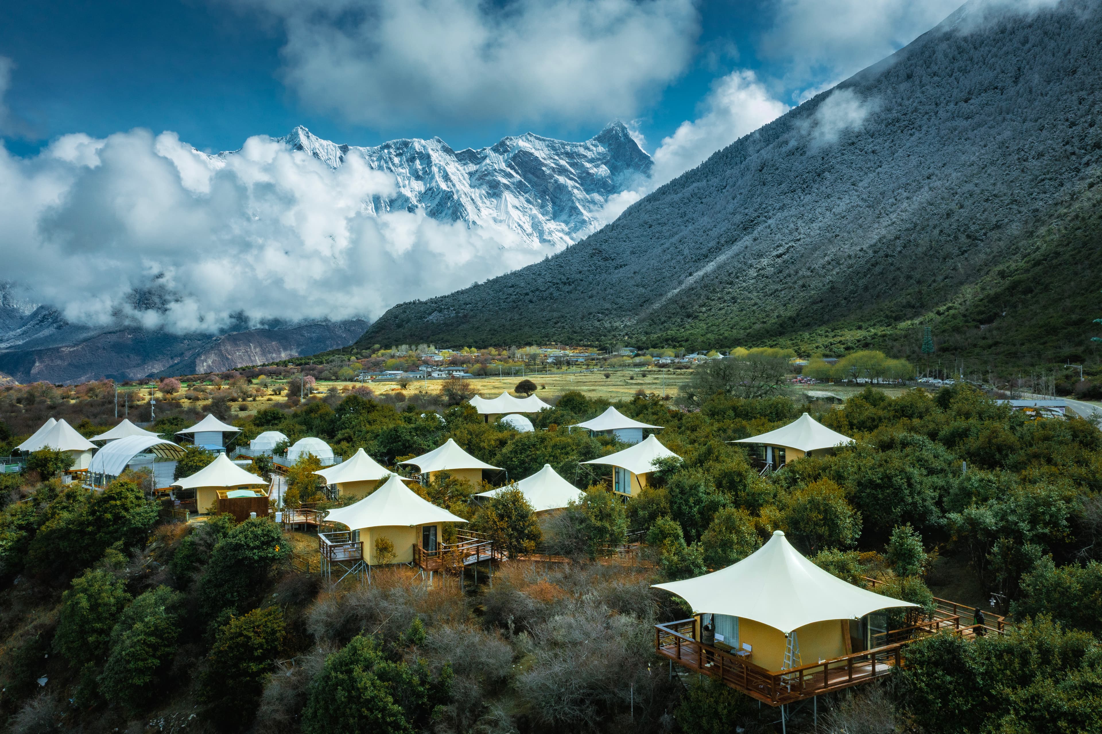 White tented cabins in a green valley below dramatic, cloud-shrouded, snow-capped mountain peaks.