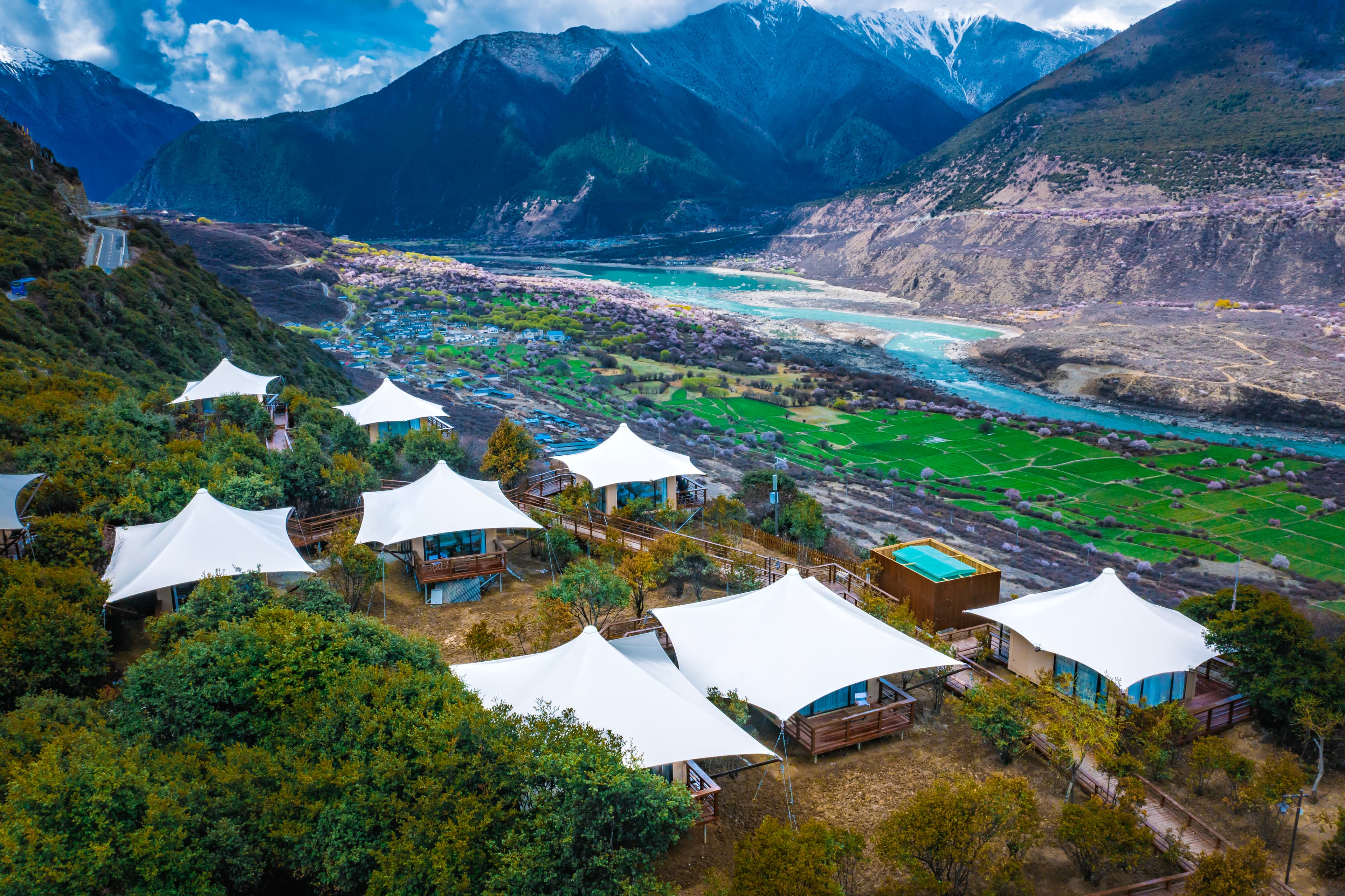 White glamping tents on a hillside overlooking a turquoise river valley and snow-capped mountains.