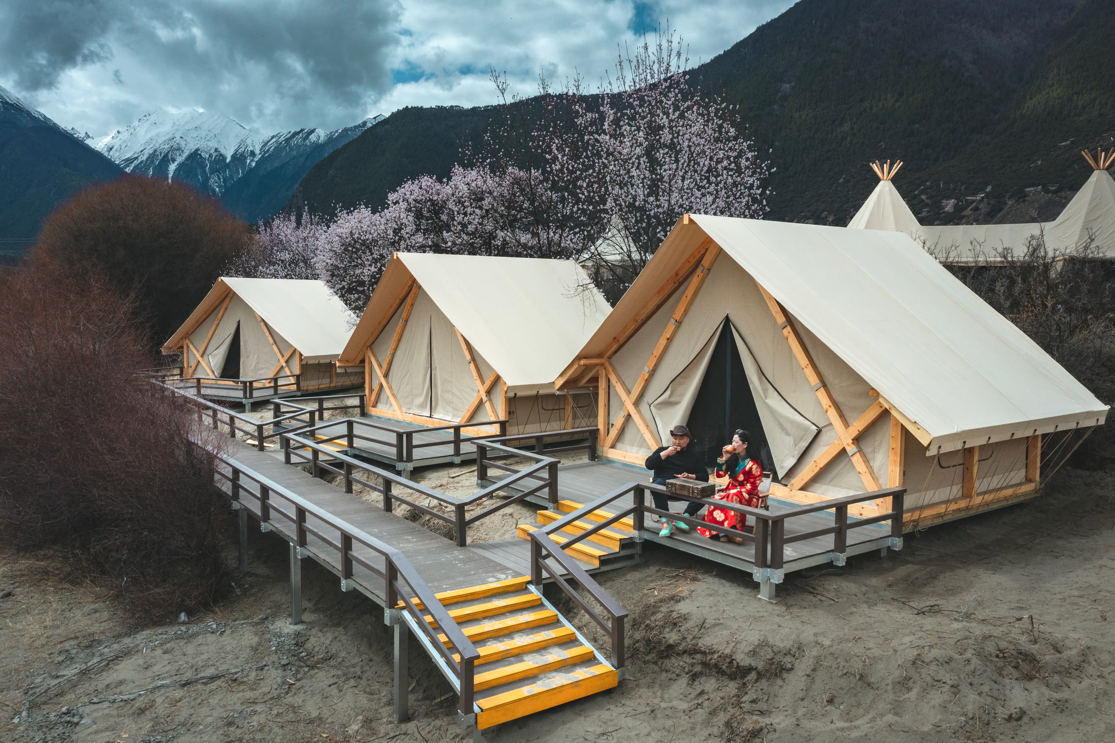 White glamping tents on wooden platforms set against snow-capped mountains and blooming cherry trees.