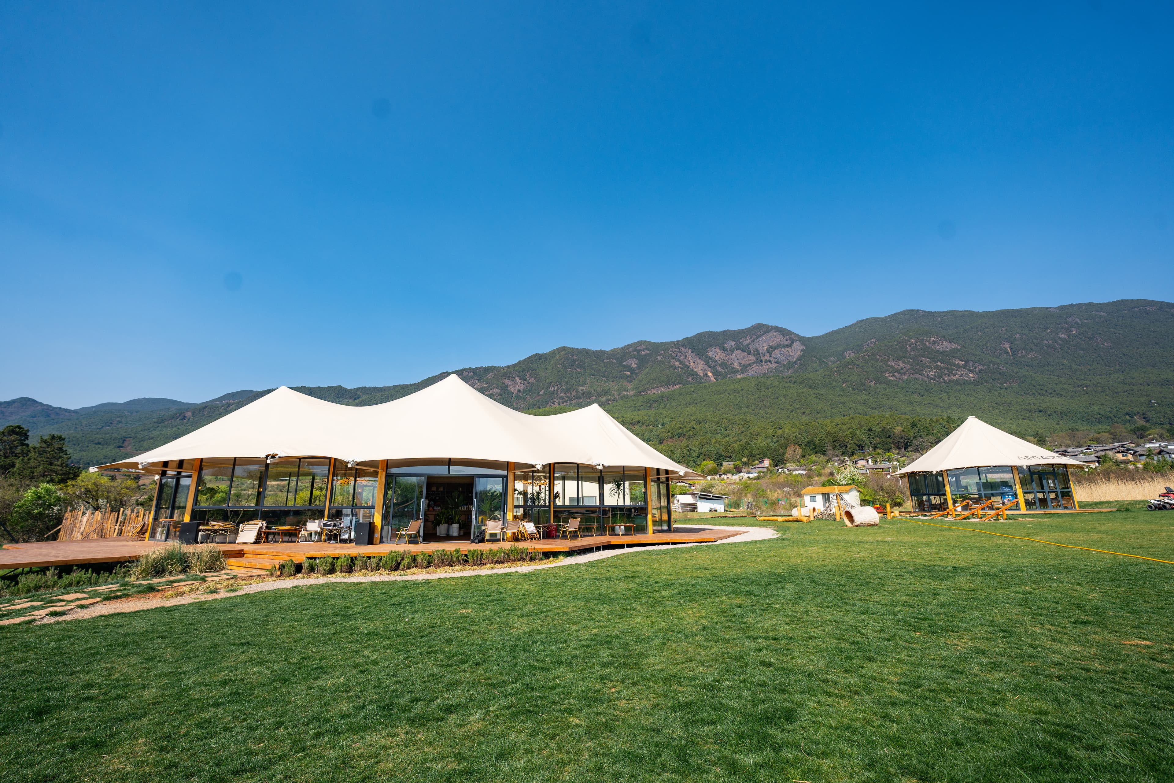 Large white tent pavilions with glass walls on a green field before forested mountains.