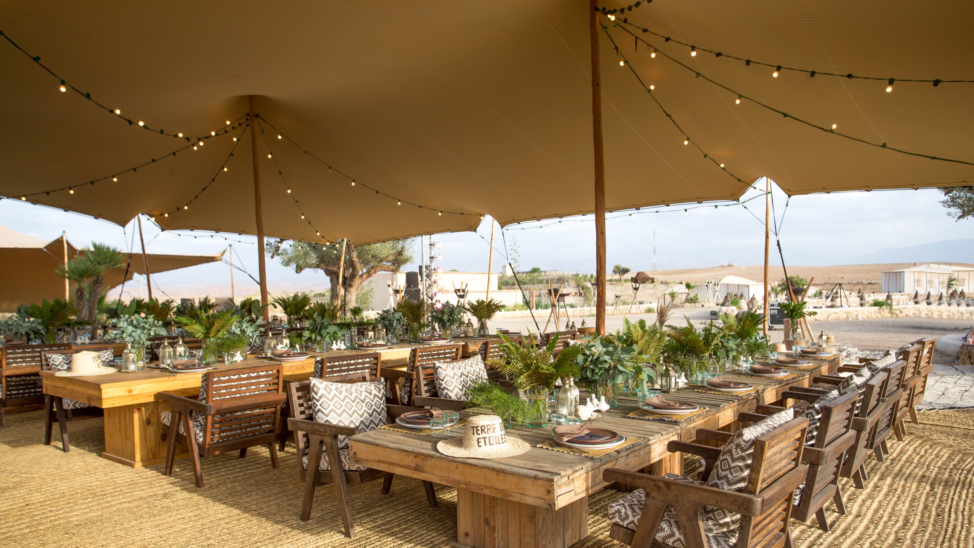 Long wooden tables with greenery centerpieces under a large tan tent decorated with string lights.
